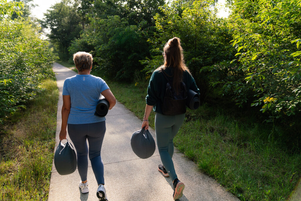 Twee vrouwen lopen met yogamat en bolster door de natuur op weg naar yoga in de natuur op de Veluwe.